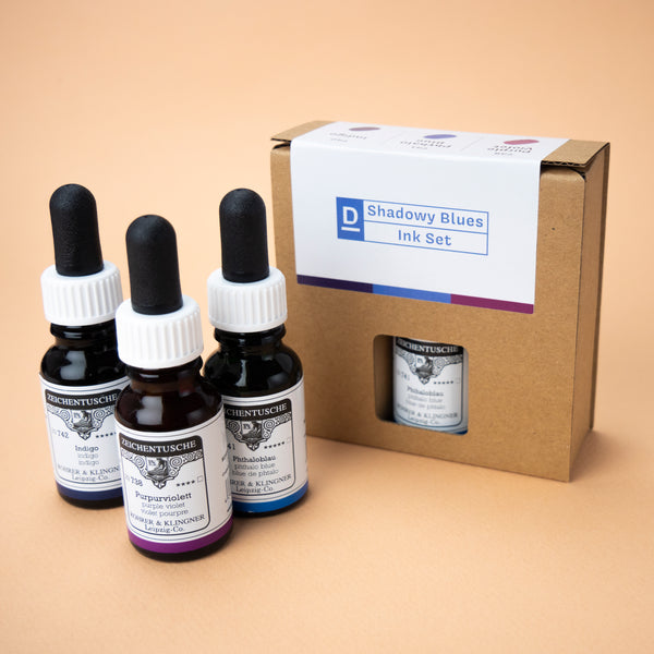 A photo of the Shadowy Blues ink set in its box and all three bottles included in the set next to it. The box has a small hole in the middle, through which the label of the color phthalo blue can be seen.
