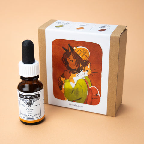 A photo of the Bark Browns ink set in its box, with one bottle of the color ochre next to it. The label on the box is a painting of a character with cat ears and curly hair, holding a cat.