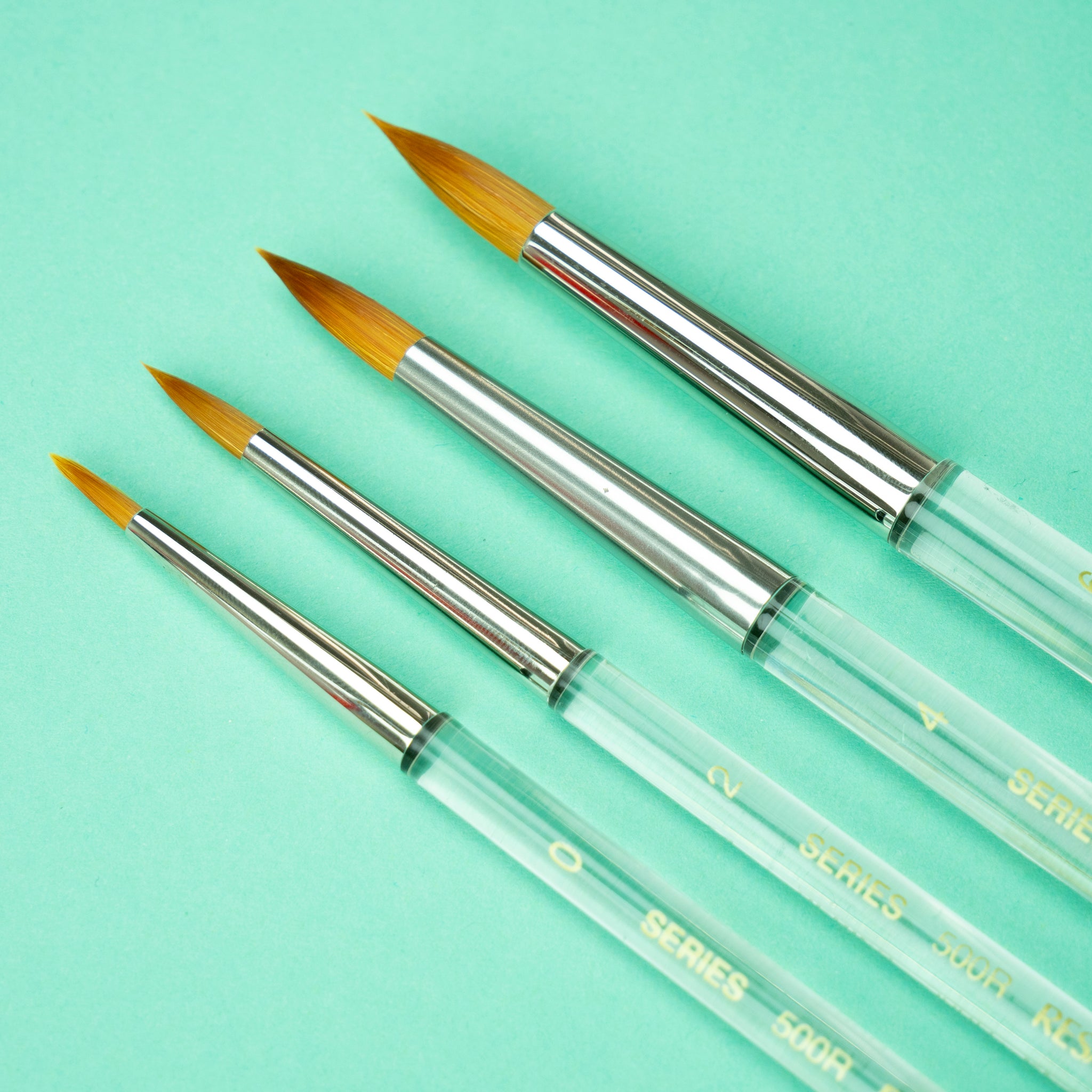 A photo of the four available brush sizes in a row on a light turquoise background, with the smallest size 0 on the left and the largest size 6 on the right. 