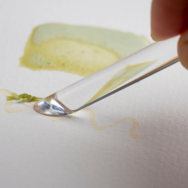 Closeup photograph of a clear plastic burnishing tool being to used to scrape dried masking fluid off of watercolor paper.